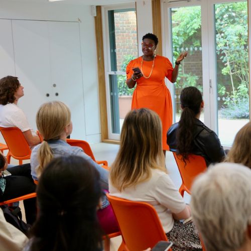 A black woman in an oramge dress stands addressing a room of people listening to her.