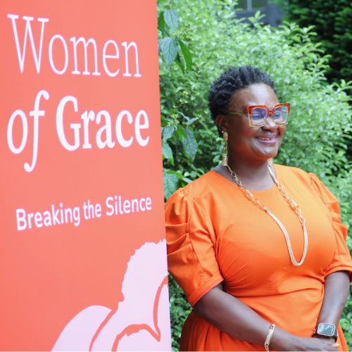 A black woman in orange dress stands nect to a banner which says Women Of Grace