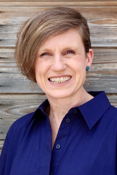Teresa Wilson is a white woman with light brown hair wearing a blue top. She is smiling into camera, standing against a slatted wooden background.