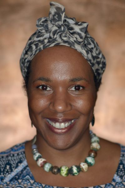 Ruth is a black woman wearing a headscarf and necklace of colourful stones. She stands in front of a golden glowing background and has a beaming smile