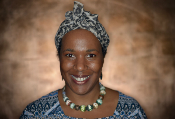 Ruth is a black woman wearing a blue head scarf. She is smiling against a golden background.